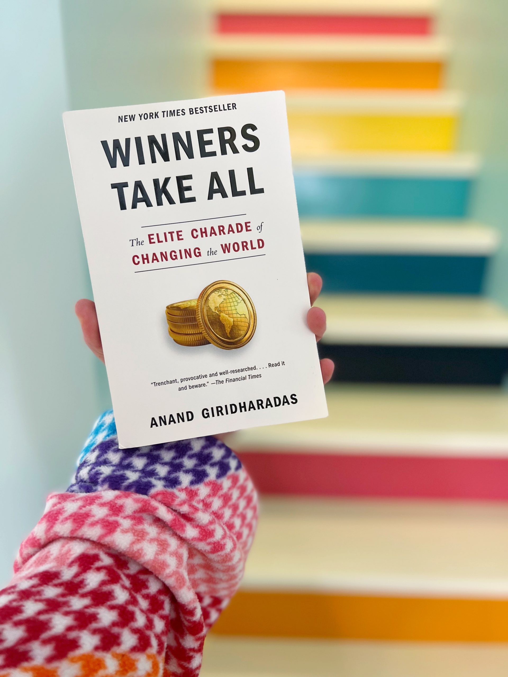 RKA holds the book "Winners Tkae All - The Elite Charade of Changing the World" by Anand Giridharadas in front of her rainbow stairs