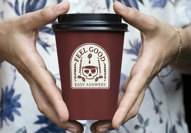 White hands holding coffee to-go cup that with skull logo that reads "Feel good easy answers"